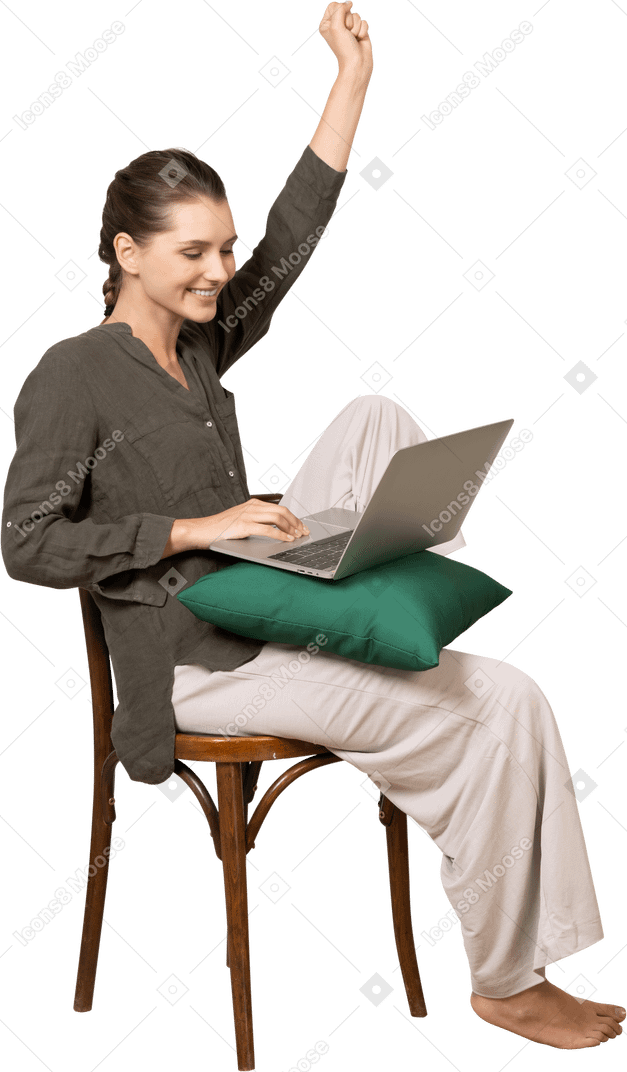 Vista di tre quarti di una giovane donna che indossa abiti da casa seduta su una sedia con un laptop e alzando la mano
