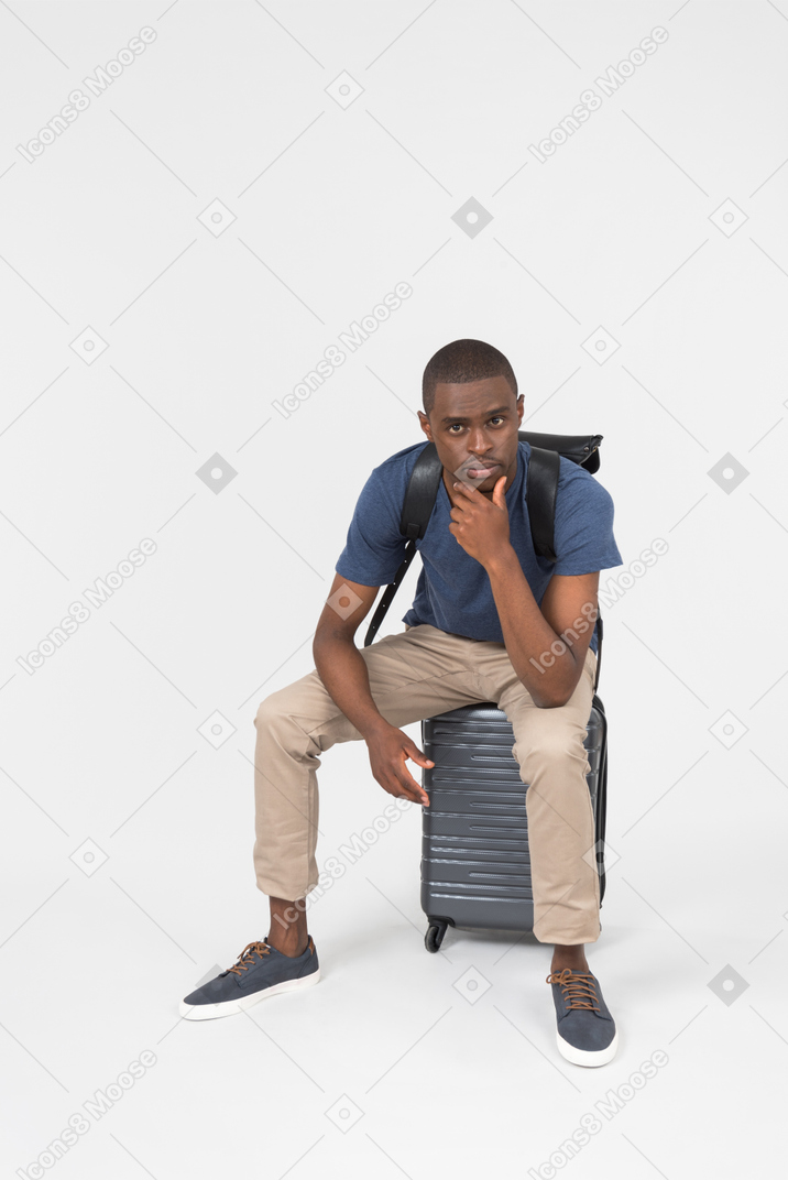 Pensive male tourist sitting on grey luggage