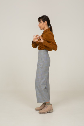 Vista lateral de uma jovem mulher asiática de calça e blusa mostrando um gesto de coração