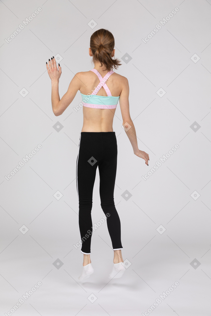 Vista posterior de una jovencita en ropa deportiva saltando y agitando la mano