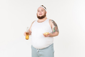 Um homem gordo no sportswear com uma garrafa de cerveja e um punhado de batatas fritas