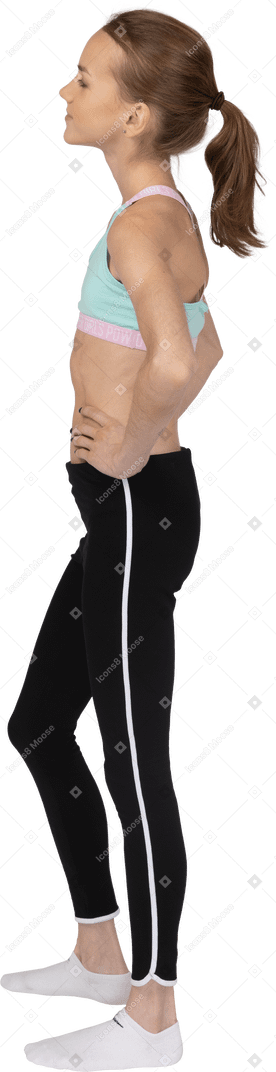 Vista laterale di una ragazza adolescente carina in abbigliamento sportivo mettendo le mani sui fianchi e guardando da parte