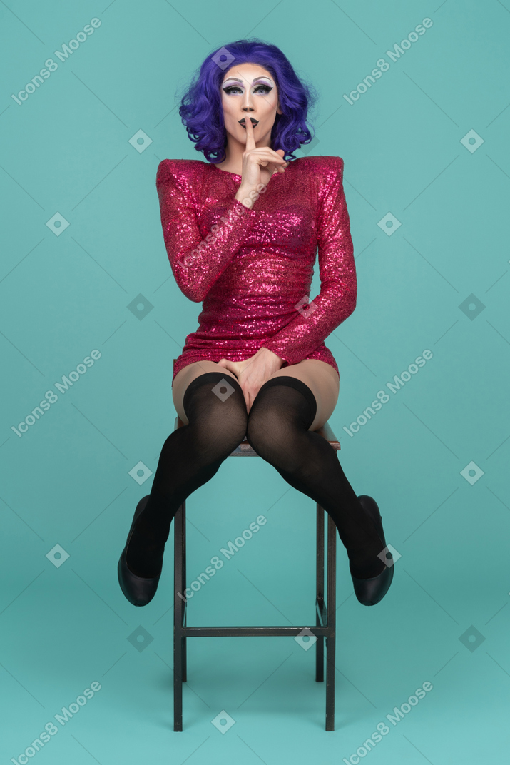 Drag queen sitting on a stool & showing silence gesture