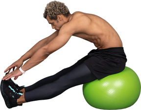 一个赤膊的黑人男子坐在绿色健身球上伸展的侧视图