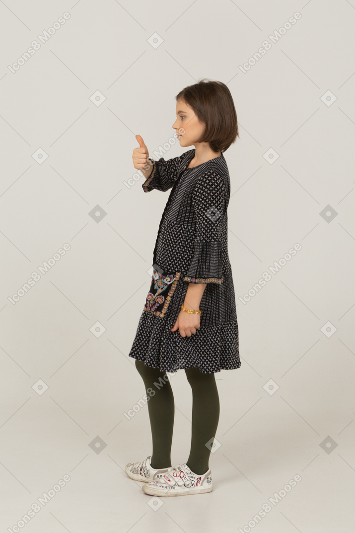 Вид сбоку маленькой девочки в платье, смотрящей в камеру и показывающей большой палец вверх