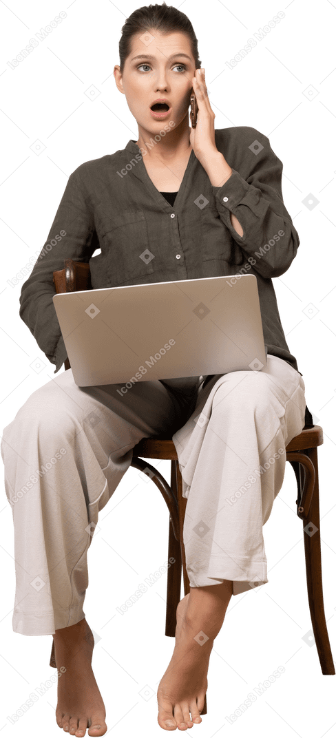 一位震惊的年轻女子坐在椅子上，拿着笔记本电脑和手机的正面视图