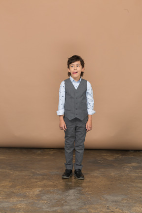 Vista frontal de um menino fofo em um terno cinza olhando para o lado e mostrando a língua