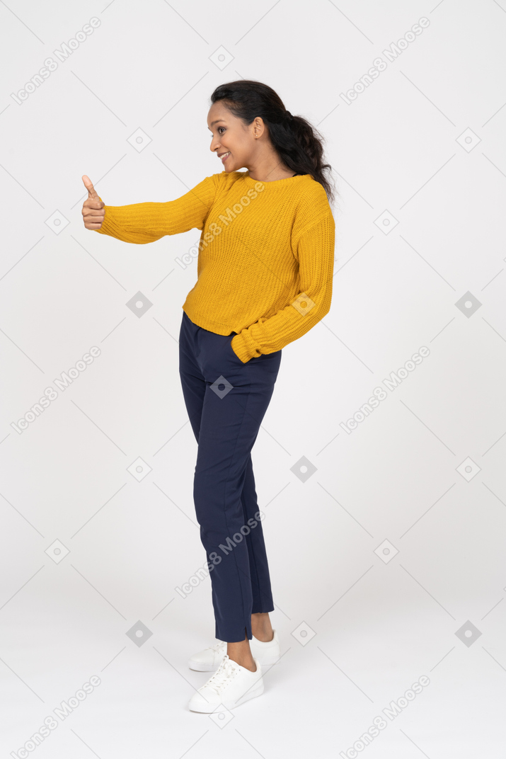Side view of a happy girl in casual clothes standing with hand in pocket and showing thumb up