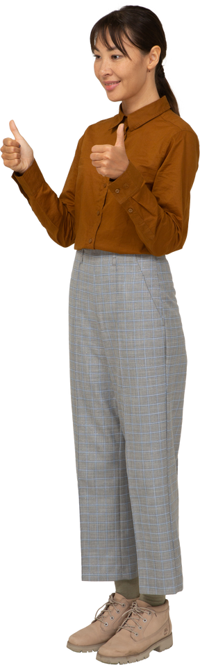 Vista di tre quarti di una giovane donna asiatica in calzoni e camicetta che mostra i pollici in su