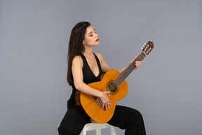 Three-quarter view of a sitting young lady in black suit holding the guitar with her eyes closed