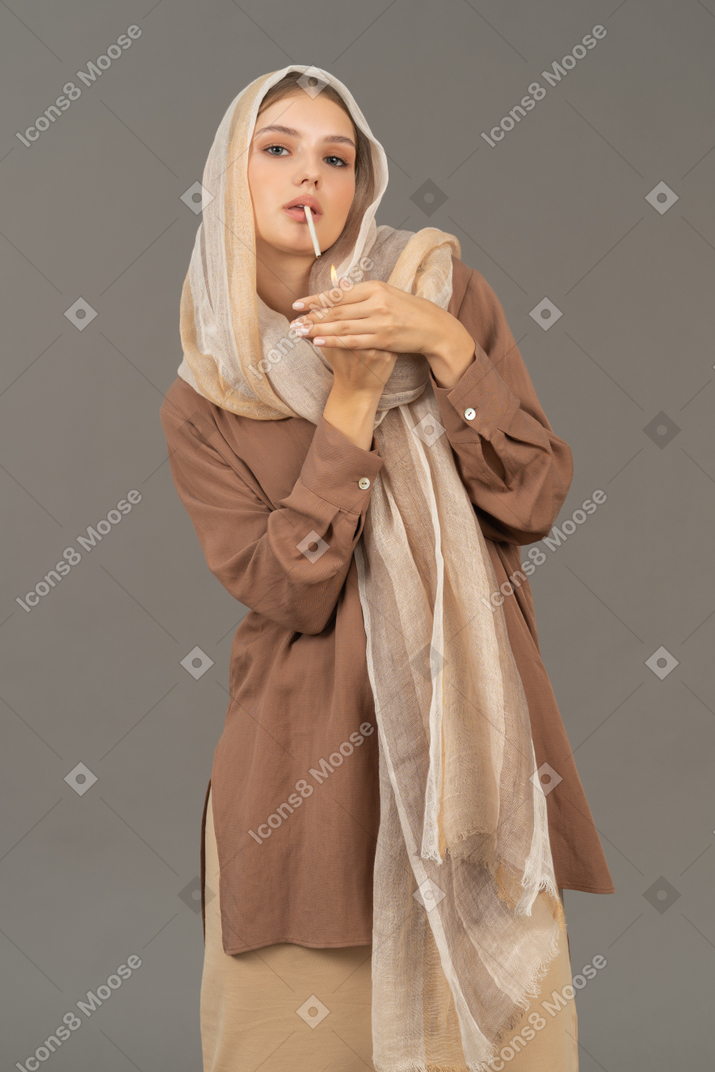 Young woman in headscarf and beige clothes lighting a cigarette