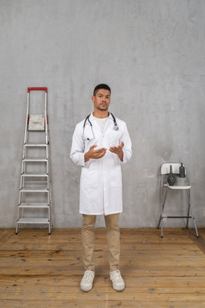 Vista frontal de un joven médico de pie en una habitación con escalera y silla explicando algo