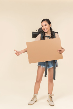 Young female hitchhiker holding paper card and showing thumb up