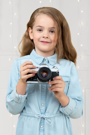 A little girl with a camera standing in front of a wall with christmas lights