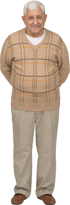 Front view of a happy old man in casual clothes standing with hands behind back and looking at camera