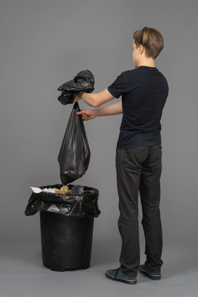 A young man holding a trash bag above a waste bin