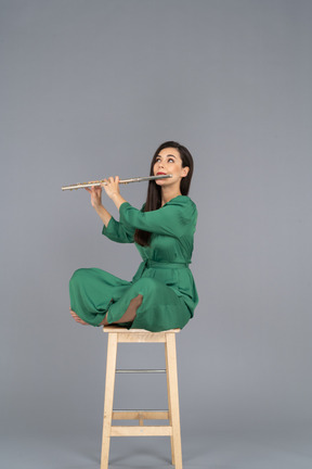 Toute la longueur d'une jeune femme jouant de la clarinette assise, les jambes croisées sur une chaise en bois