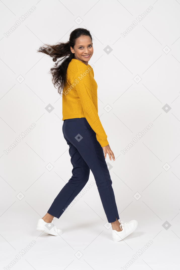 Vista lateral de una niña feliz en ropa casual caminando y mirando a la cámara