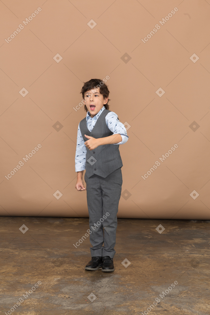 Вид спереди на симпатичного мальчика в сером костюме, показывающего большой палец вверх