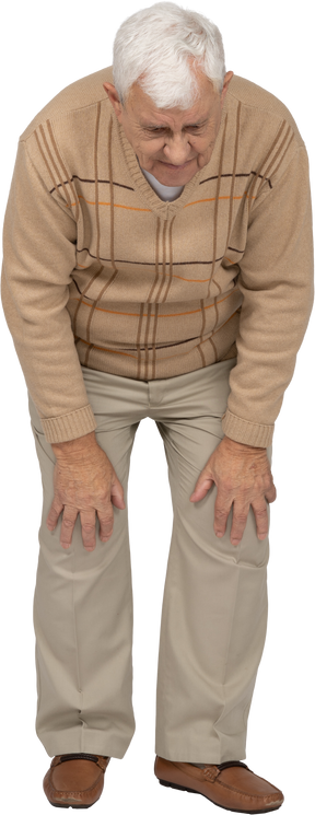 Front view of an old man in casual clothes bending down and touching his hurting knees