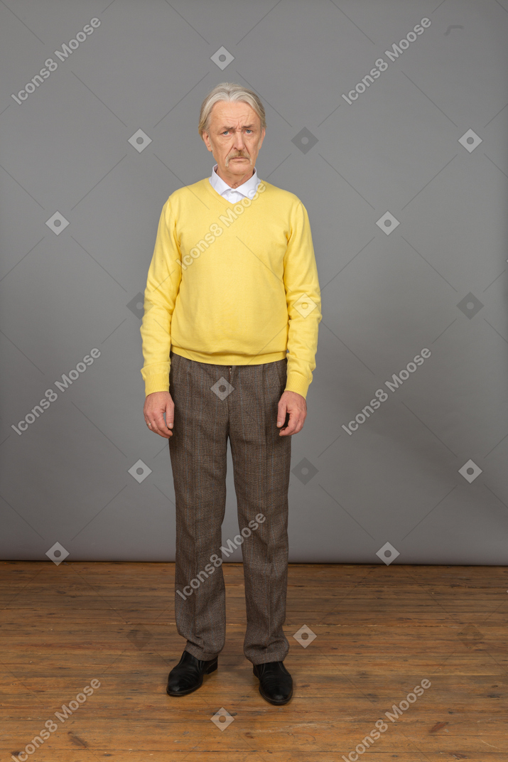 Front view of an old displeased man in yellow pullover knitting brows and looking at camera
