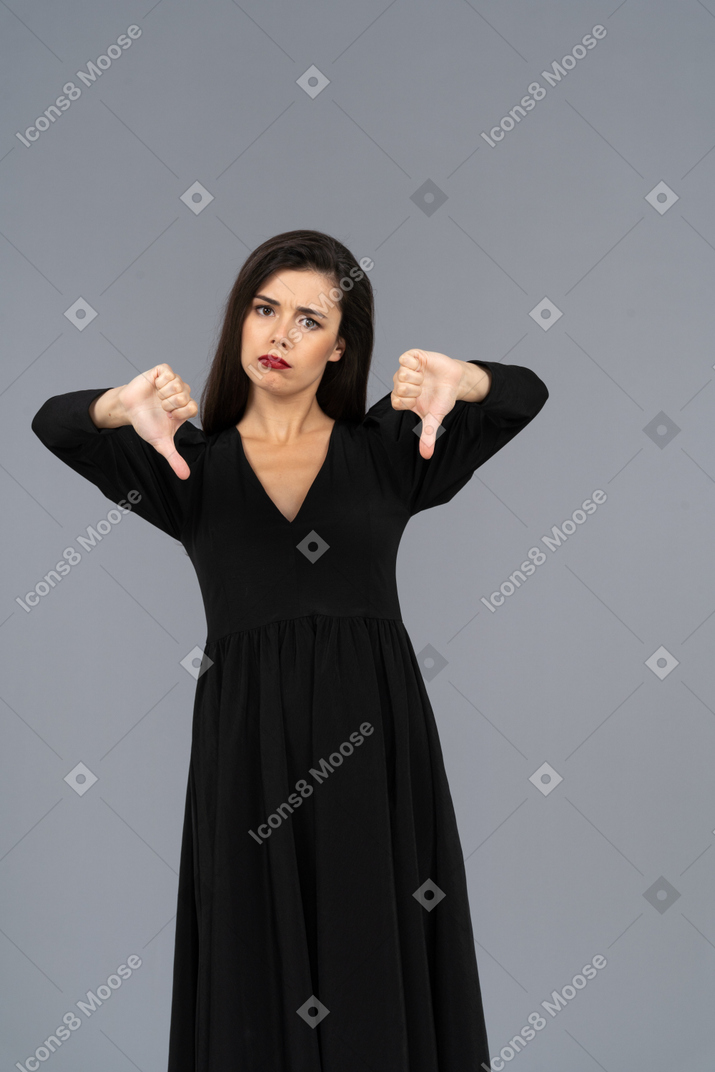 Vista frontal de uma jovem insatisfeita de vestido preto com os polegares para baixo