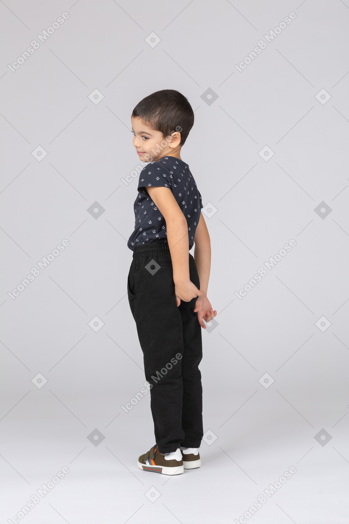 Vista lateral de un niño tímido en ropa casual de pie con las manos detrás de la espalda y mirando hacia abajo