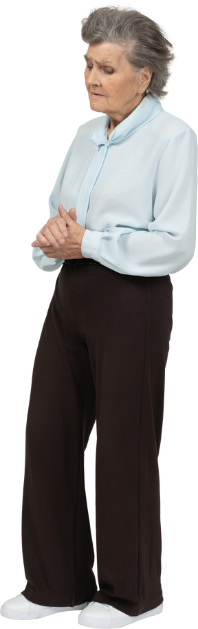 Три четверти вида недовольной пожилой женщины в блузке и брюках, держащей руки вместе и смотрящей вниз