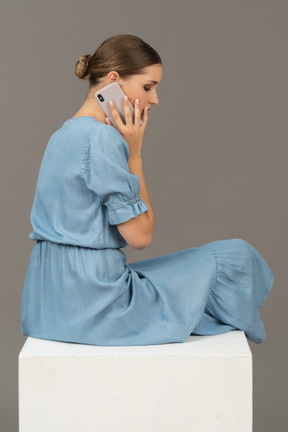 Side-back view of young woman sitting on cube and talking on smartphone