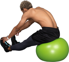 Vista posteriore di tre quarti di un uomo afro a torso nudo che si allunga mentre è seduto su una palla verde da palestra