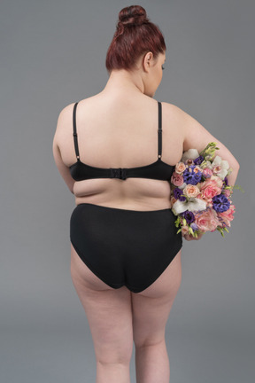 Cuerpo femenino positivo en lencería negra posando de espaldas a la cámara con un ramo de flores