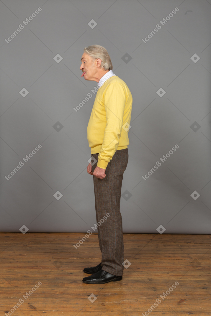 Vista laterale di un vecchio uomo curioso in pullover giallo che mostra la lingua e guardando da parte