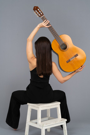 Back view of a young lady in black suit holding the guitar over head and sitting on stool