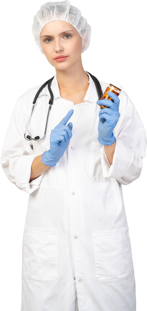 Front view of a young female doctor pointing finger at the jar of pills