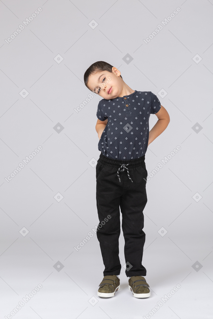 Vue de face d'un garçon mignon dans des vêtements décontractés posant avec les mains sur le dos et regardant la caméra
