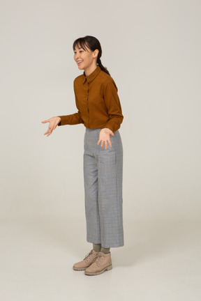 Vista di tre quarti di una giovane donna asiatica gesticolante sorridente in calzoni e camicetta