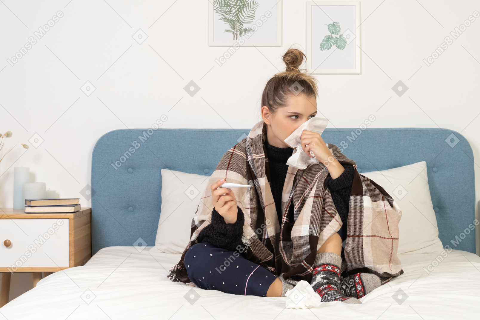 Vista frontal de una joven enferma en pijama envuelto en una manta de cuadros sonándose la nariz