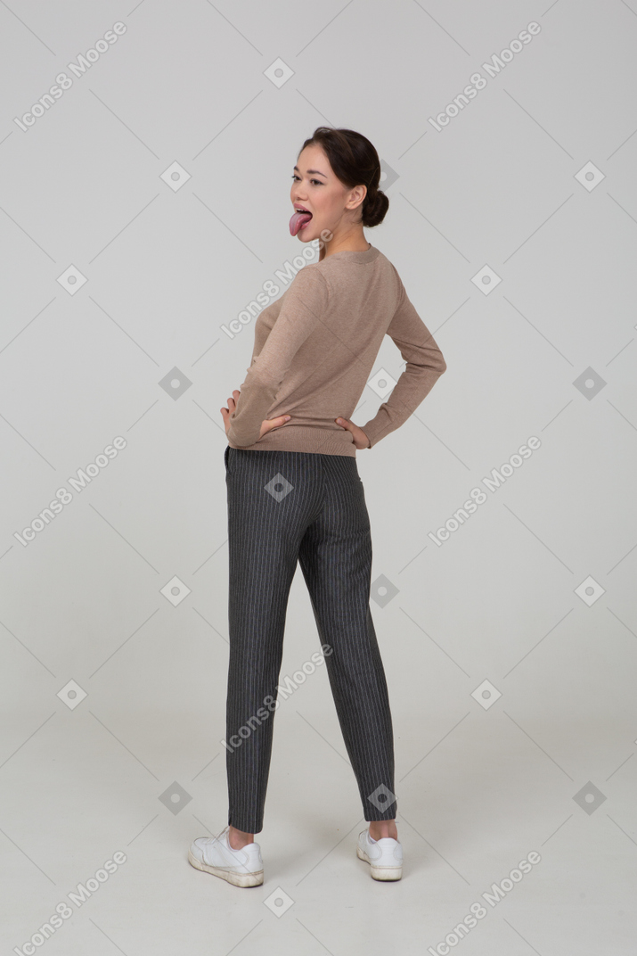 Three-quarter back view of a young lady in pullover and pants putting hands on hips and showing tongue