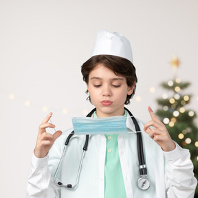 A little boy in doctor's uniform putting on a face mask with a christmas tree on background