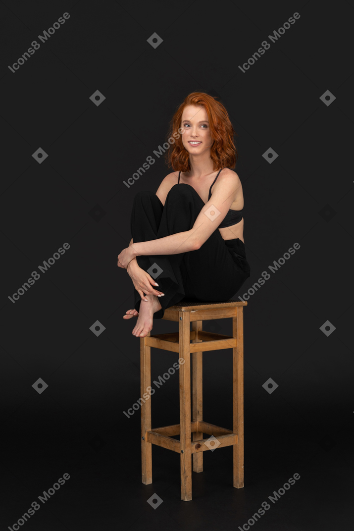 검은 바지와 브래지어를 입은 아름다운 여인의 3/4 정면보기, 나무 의자에 앉아 무릎을 껴안기