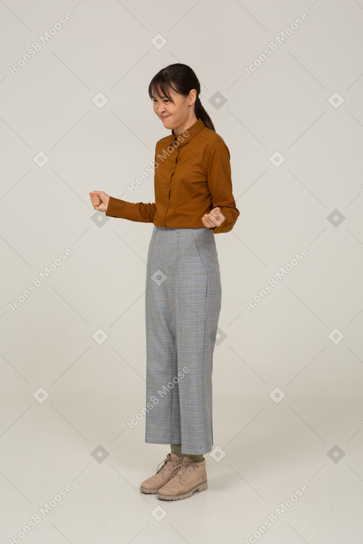 Vista di tre quarti di una giovane donna asiatica malvagia in calzoni e camicetta che stringe i pugni