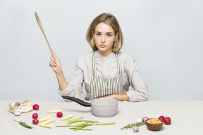 Chica en delantal sosteniendo una cuchara de madera y sentada en la mesa con pan y verduras en ella