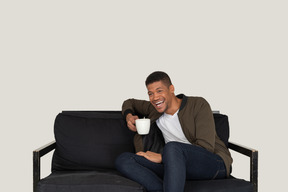 一个笑着的年轻人坐在沙发上，拿着一杯咖啡的前视图