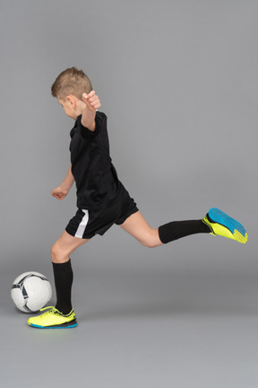 Vista lateral de un niño chico en uniforme de fútbol pateando una pelota