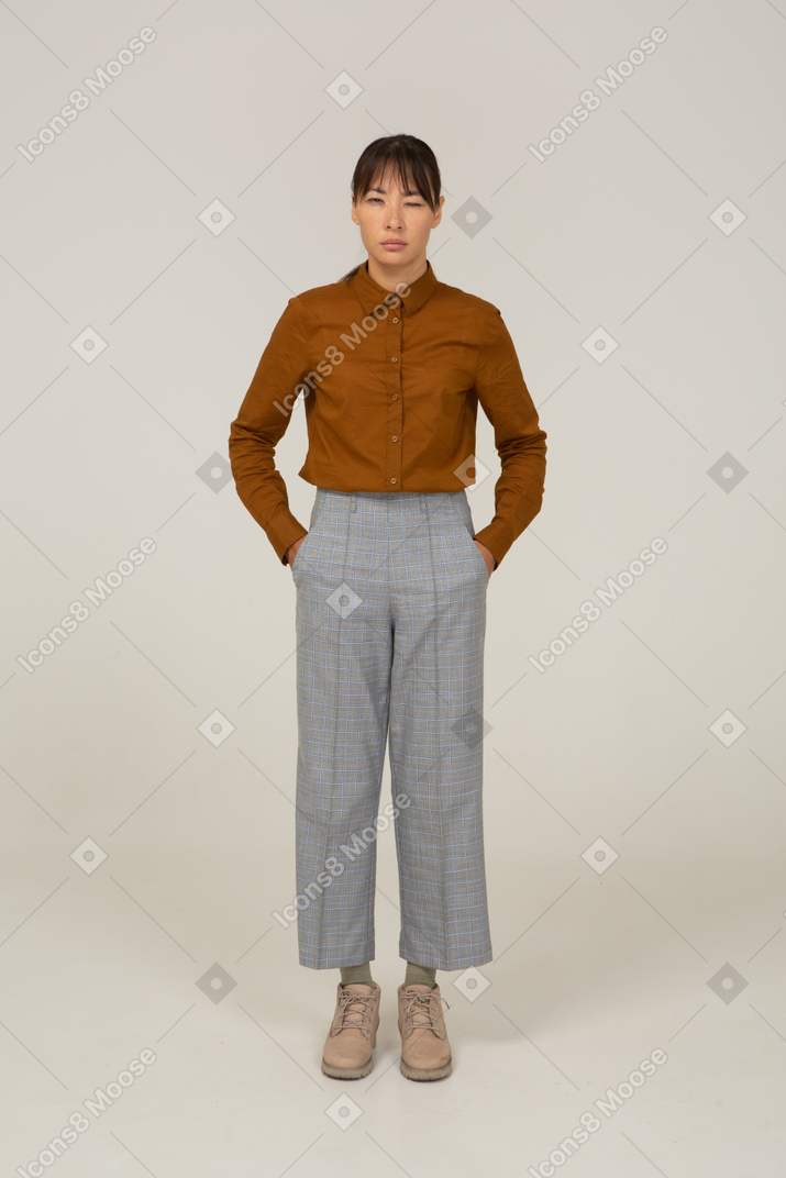Vista frontal de uma jovem mulher asiática de calça e blusa estreitando os olhos e colocando as mãos nos bolsos