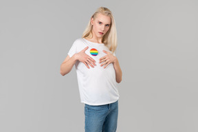 Beau jeune homme aux longs cheveux blonds, debout sur un fond gris, vêtu d'un jean bleu et d'un t-shirt blanc avec écusson lgbt