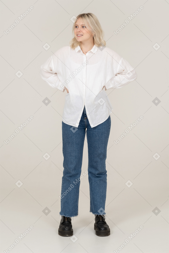 Vue de face d'une femme blonde dans des vêtements décontractés mettant les mains sur les hanches et regardant de côté en souriant