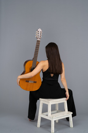 Back view of a young lady in black suit holding the guitar and sitting on stool