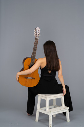 Vue arrière d'une jeune femme en costume noir tenant la guitare et assis sur un tabouret