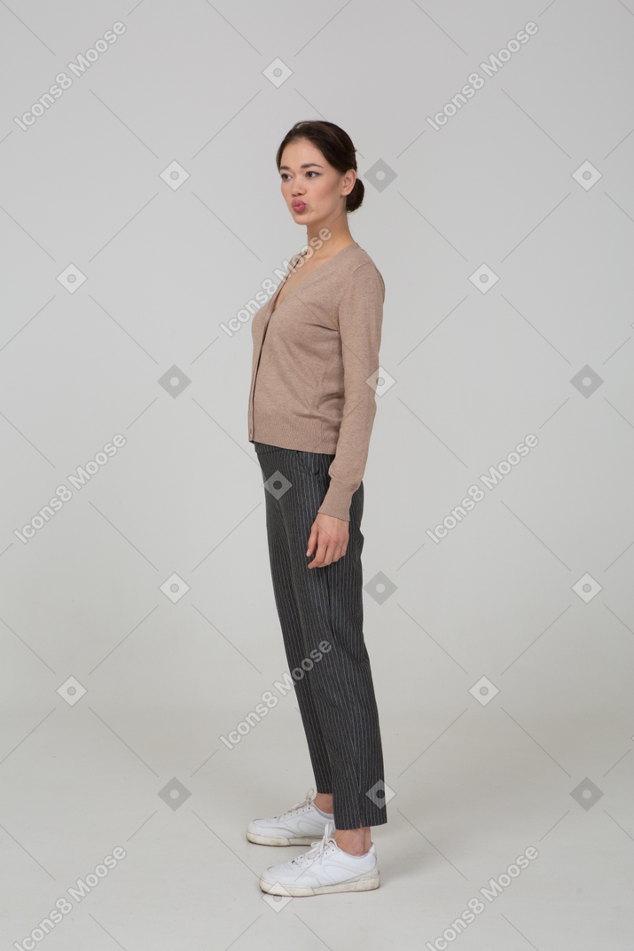 Vista di tre quarti di una giovane donna in piedi ancora in pullover e pantaloni mettendo la mano sui fianchi e imbronciato
